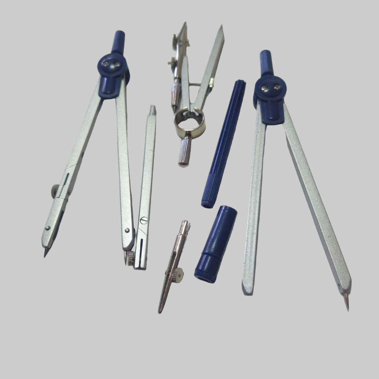 A set of metal compasses and drawing tools with blue plastic knobs and covers, arranged on a plain off-white surface.